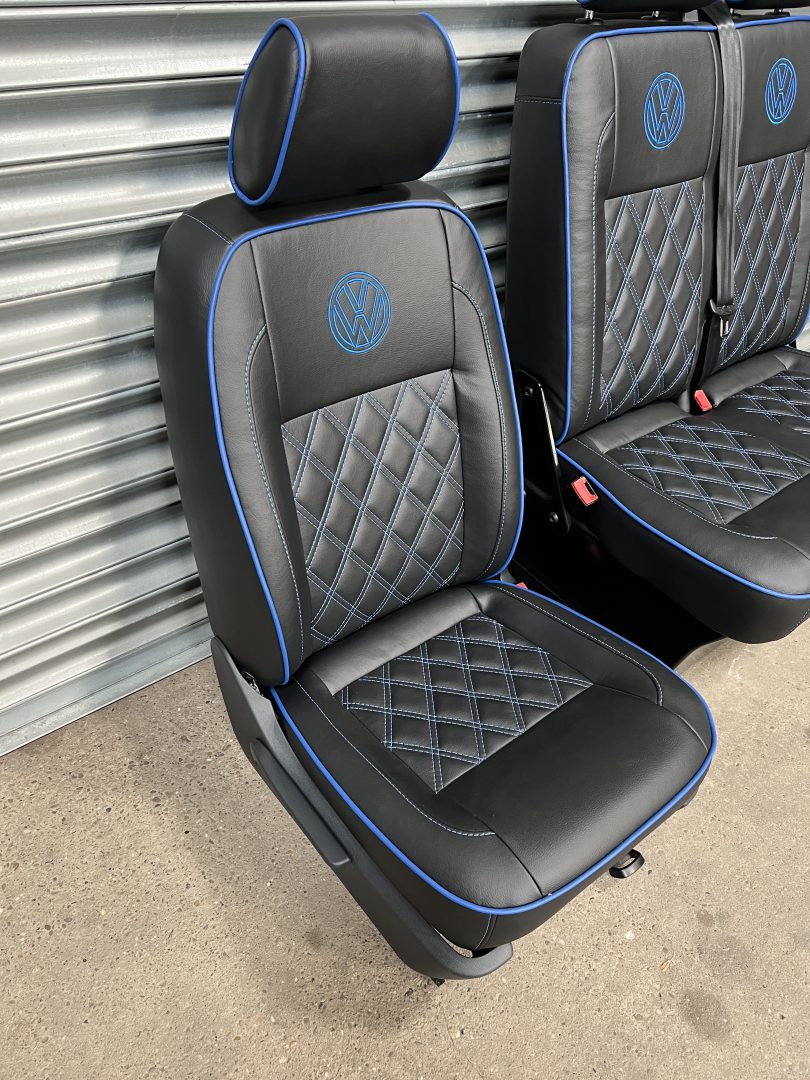 VW T5 T6 front seats, angled view, highlighting the driver seat in black leatherette with blue cross stitching, piping and VW logo.