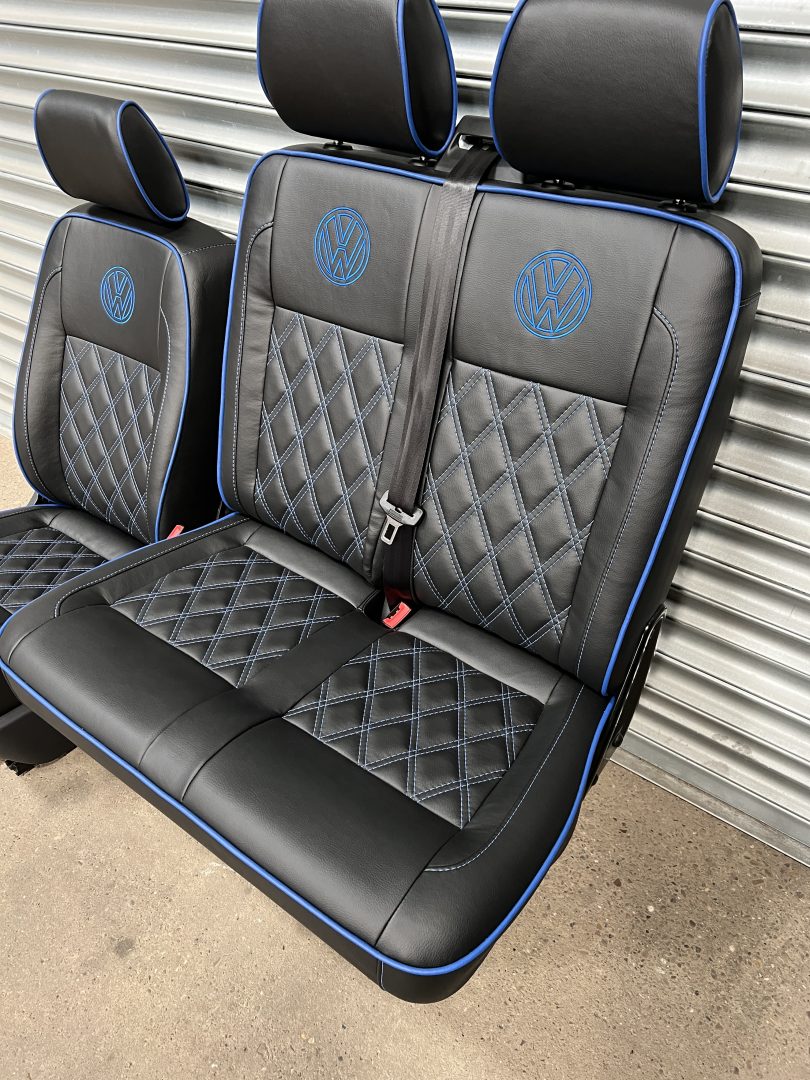 VW T5 T6 front seats, angled view, highlighting the double seat in black leatherette upholstery with blue cross stitching, piping and VW logos.